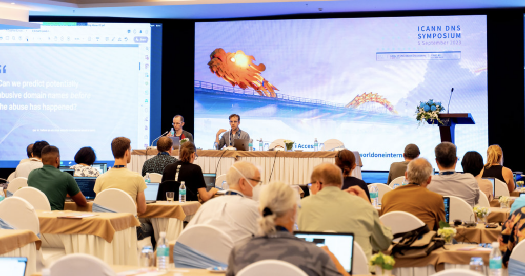 People attending a session in a conference room during the ICANN DNS Symposium 2019. The main screen displays a presentation slide, and two speakers are seated at the front table.